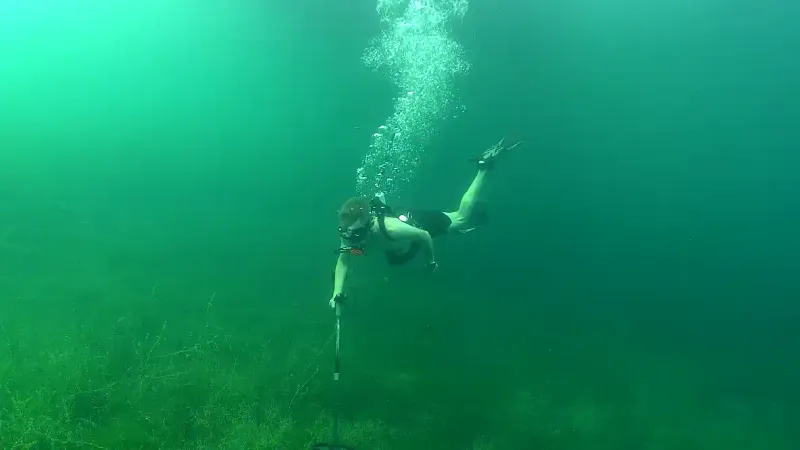 Ein Taucher schwimmt mit einem Metalldetektor unter Wasser, während über ihm Luftblasen aufsteigen und grüne Wasserpflanzen den Seegrund bedecken.