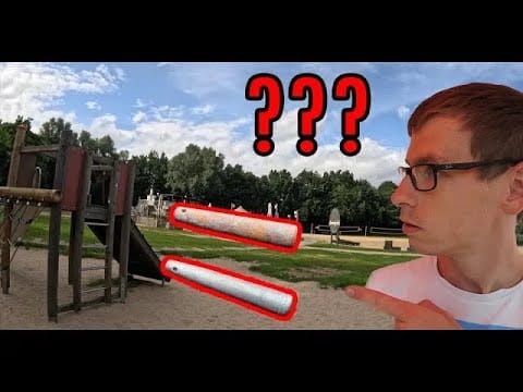 Ein Mann mit Brille zeigt auf zwei rot umrandete Metallstangen auf einem Spielplatz, über ihm stehen drei große Fragezeichen - ein Hinweis auf eine spannende Schatzsuche metalldetektor Szene inmitten von Bäumen und Spielgeräten im Freien.