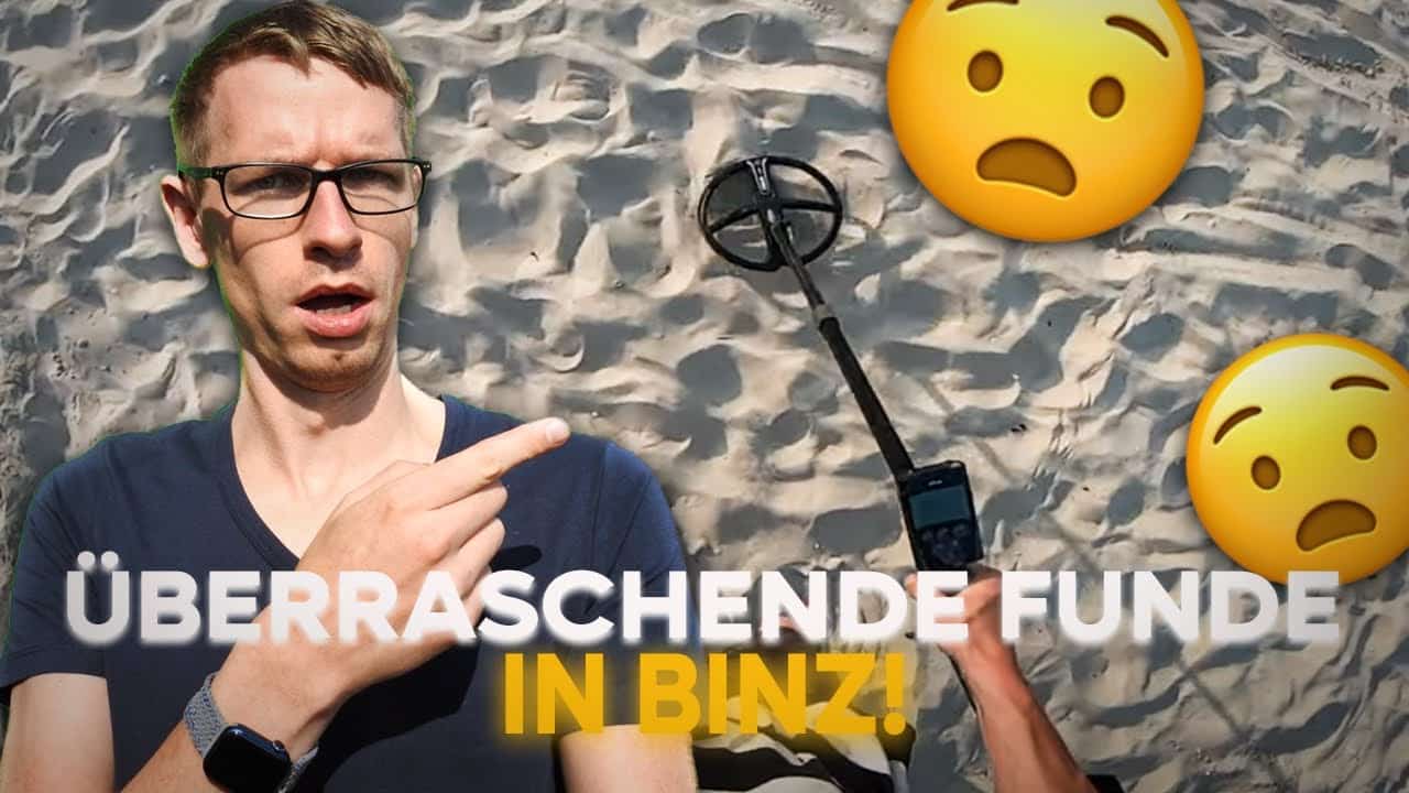 Ein überraschter Mann zeigt auf Sand, während er einen Metalldetektor in der Hand hält. Zwei schockierte Gesichts-Emojis sind über ihm zu sehen. Der Text auf Deutsch lautet "ÜBERRASCHENDE FUNDE IN BINZ!" - vielleicht ein Ring verloren gefunden oder sogar ein Handy im Wasser verloren!.