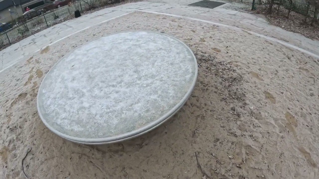 Ein großer, runder Spielplatzspinner aus Metall liegt auf sandigem Boden, seine frostige Oberfläche deutet auf einen kühlen Morgen hin. Rundherum sind Fußabdrücke zu sehen - vielleicht von jemandem, der in der Nähe ein Abenteuer auf der Schatzsuche metalldetektor erlebt hat. Im Hintergrund sind ein eingezäuntes Gelände und gepflasterte Wege zu sehen.