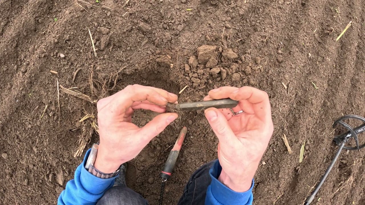 Eine Person in einem blauen Sweatshirt hält einen kleinen, schlammigen Metallgegenstand - möglicherweise einen im Garten gefundenen Ring - in der Hand, während in der Nähe ein Metalldetektor und ein Grabungswerkzeug auf dem Boden zu sehen sind.