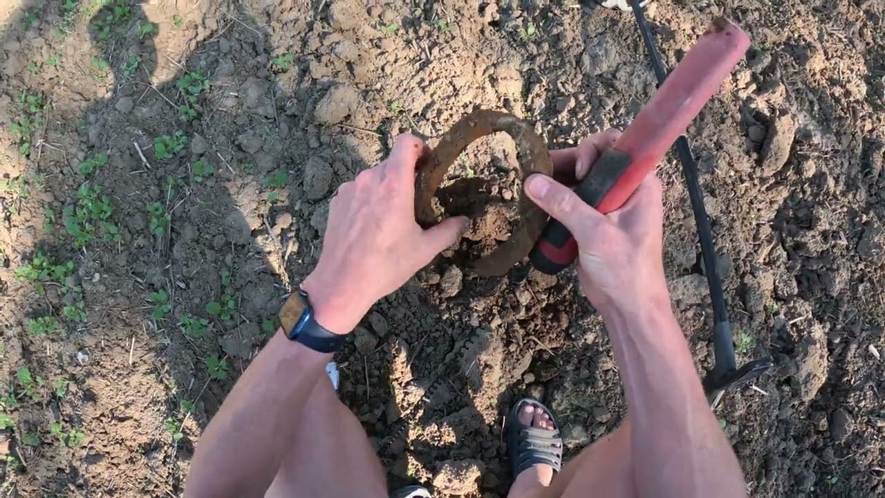 Eine Person steht auf trockenem, rissigem Boden und hält ein schlammiges Hufeisen in der einen und ein Grabwerkzeug in der anderen Hand - eindeutig inspiriert von einer Schatzsuche mit Metalldetektor. Kleine grüne Pflanzen sprenkeln den Boden; die Person trägt eine Uhr und Sandalen.