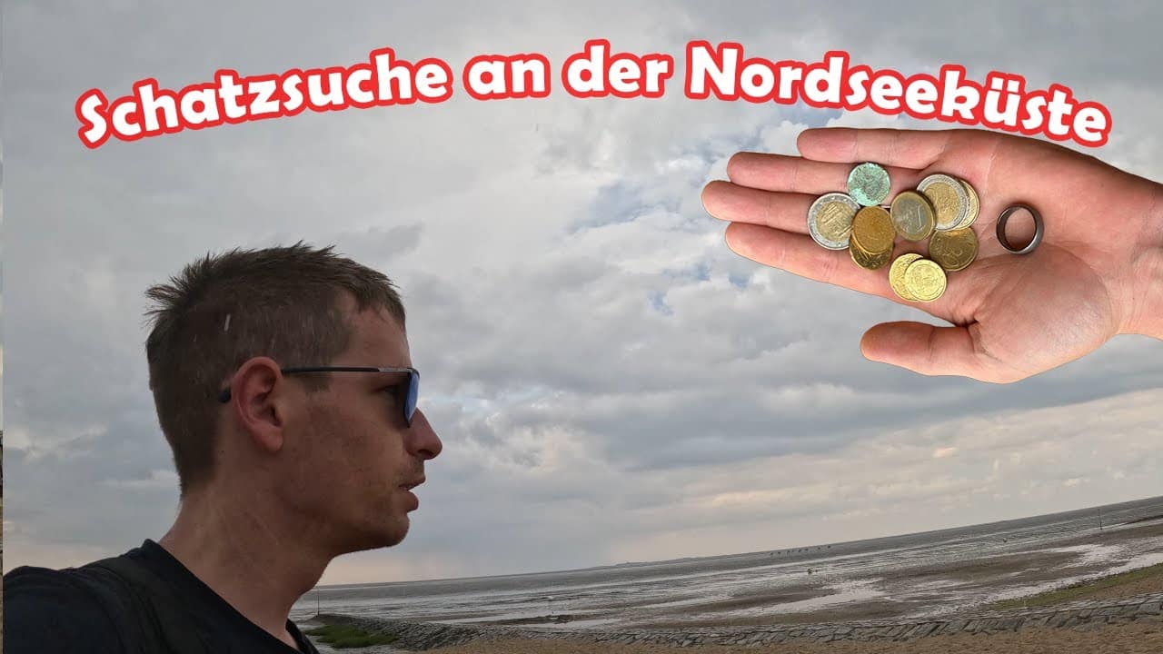 Eine Person steht an einem Strand unter einem bewölkten Himmel. Eine Hand in Großaufnahme hält verschiedene Münzen und einen Ring, Ring verloren gefunden. Der deutsche Text lautet: "Schatzsuche an der Nordseeküste".
