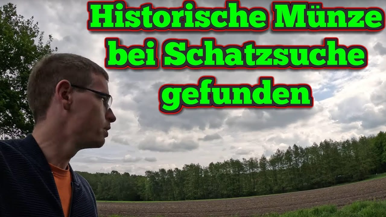 Eine Person mit kurzen Haaren und Brille steht vor einem gepflügten Feld und blickt in die Ferne. Der Text auf Deutsch lautet: Historische Münze bei Schatzsuche metalldetektor gefunden in grün und rot. Der Himmel ist bewölkt.