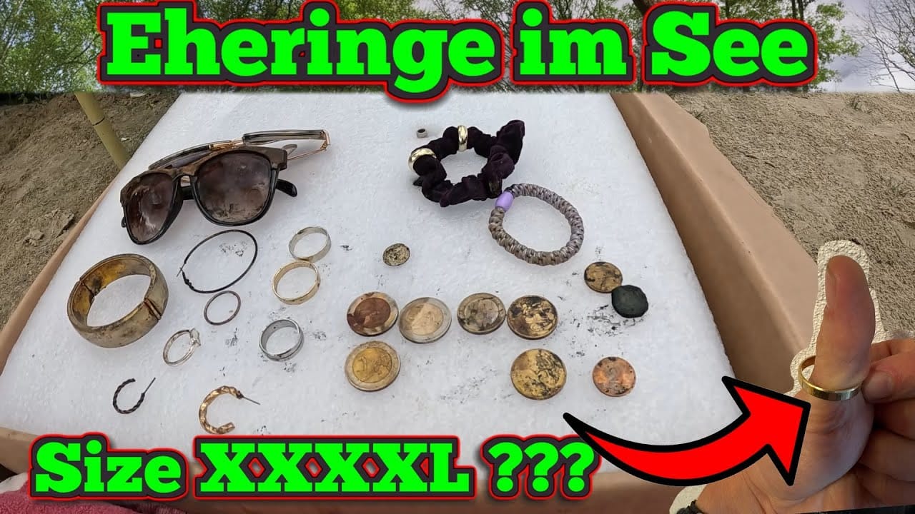 Eine Ausstellung von Schmuck, Ringen, Münzen und Sonnenbrillen auf einer weißen Tafel im Freien mit dem deutschen Text "Eheringe im See" und "Größe XXXXXL ???" deutet auf Schätze hin, die im Wasser verloren oder im See verloren sind. Eine Hand hält einen großen Ring, der durch einen roten Pfeil hervorgehoben wird.