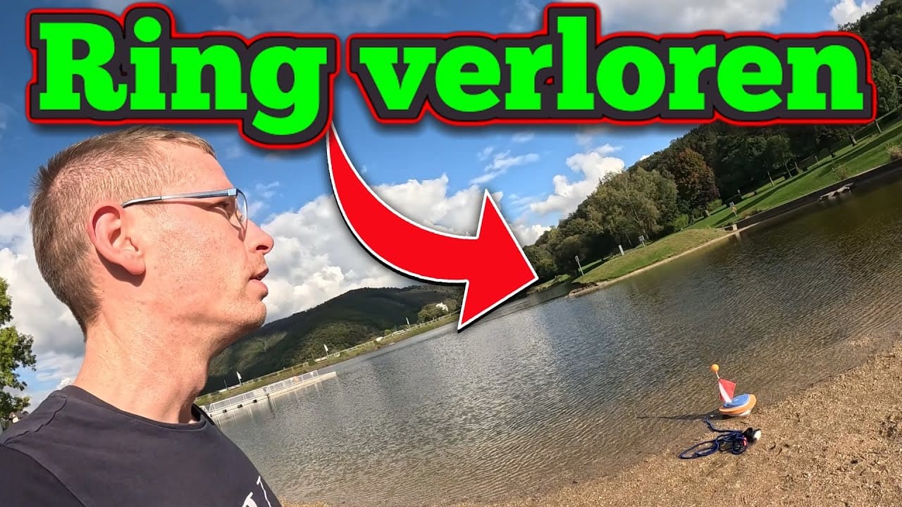 Ein Mann steht am Ufer eines Sees und blickt auf das Wasser. Darüber steht in leuchtendem Grün und Rot die Aufschrift "Ring verloren" mit einem roten Pfeil, der in Richtung See zeigt. Ein Spielzeugboot liegt am sandigen Ufer - vielleicht etwas anderes im See verloren?.