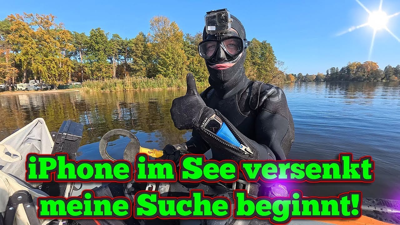 Ein Taucher mit Neoprenanzug und Schwimmbrille sitzt in einem Boot auf einem See und zeigt den Daumen nach oben. Der Text auf Deutsch lautet: iPhone im See versenkt, meine Suche beginnt! Handy im Wasser verloren? Zeit für eine Schatzsuche mit Metalldetektor!.