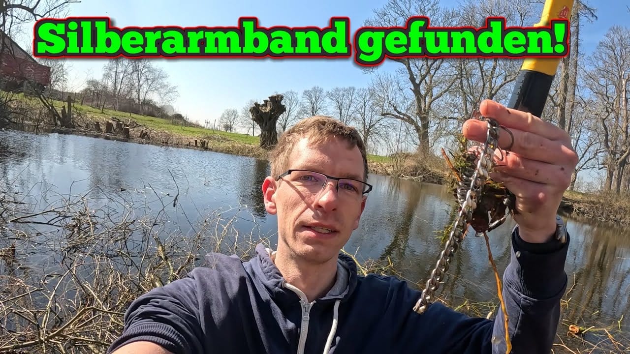 Ein Mann hält ein silbernes Armband hoch und schaut in die Kamera an einem Teich, an dem Schätze wie "Ring verloren gefunden" oder Gegenstände "im see verloren" entdeckt werden könnten. Im Hintergrund sind Bäume und Sträucher zu sehen. In leuchtendem Grün und Rot steht "Silberarmband gefunden".