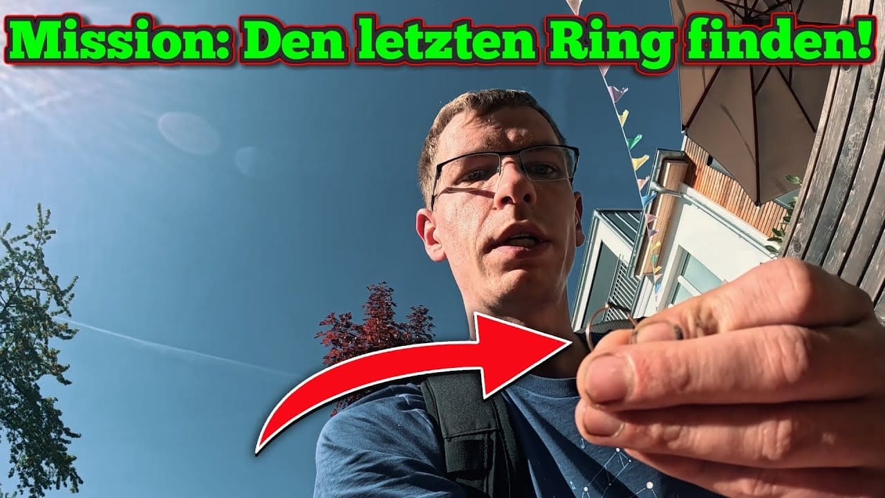 Ein Mann mit Brille hält einen kleinen Gegenstand mit einem Metallring - perfekt für eine Schatzsuche mit Metalldetektor - und blickt in die Kamera im Freien. Ein Pfeil zeigt auf den Gegenstand. Der Text darüber lautet: Mission: Den letzten Ring finden! auf Deutsch.