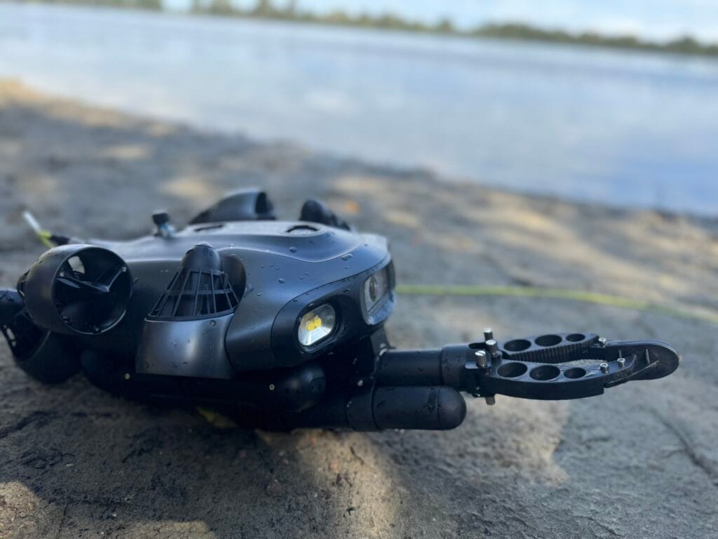 Eine schwarze Unterwasserdrohne mit einem Roboterarm ruht auf sandigem Boden in der Nähe eines Gewässers, wobei Bäume und Himmel im Hintergrund verschwimmen.