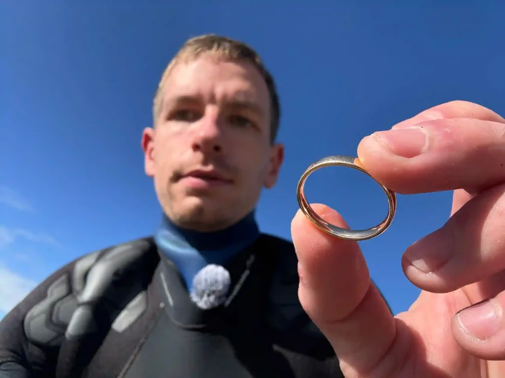 Eine Person im Neoprenanzug hält einen goldenen Ring in die Kamera, im Hintergrund ein strahlend blauer Himmel - wie eine Szene aus dem schatzsucher tv, perfekt für alle, die sich für schatzsuche tv oder Geschichten von "Ring verloren" interessieren.