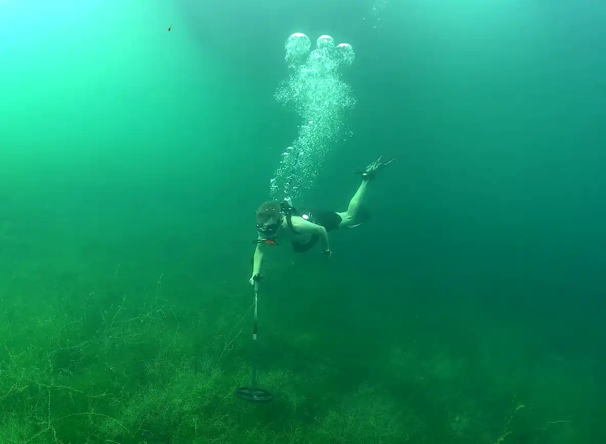 Ein Taucher schwimmt mit einem Metalldetektor unter Wasser, während über ihm Luftblasen aufsteigen und grüne Wasserpflanzen den Seegrund bedecken.